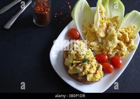 Scrambled eggs with spices and herbs on crunchy whole grain toast and vegetables in a white plate on a black background. Healthy breakfast. Top view. Stock Photo