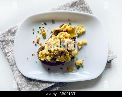 Scrambled eggs with spices and herbs on crunchy whole grain toast in a white plate on a white wooden background. Healthy breakfast. Directly above. Stock Photo