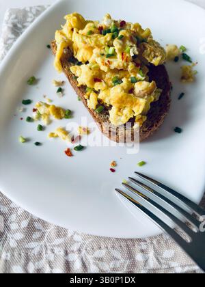 Scrambled eggs with spices and herbs on crunchy whole grain toast in a white plate on a white wooden background. Healthy breakfast. Directly above. Stock Photo
