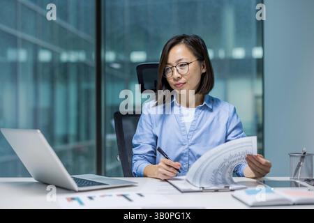 Serious confident businesswoman doing paperwork, successful financier concentrated preparing financial report. Asian accountant working at desk inside office. Stock Photo