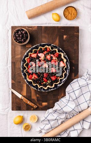 Unbaked berry pie with strawberries and blueberries in a fluted crust, set on a wooden cutting board with baking ingredients and kitchen tools. Stock Photo