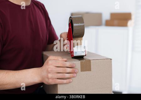 Man using dispenser with adhesive tape to seal cardboard box indoors, closeup Stock Photo