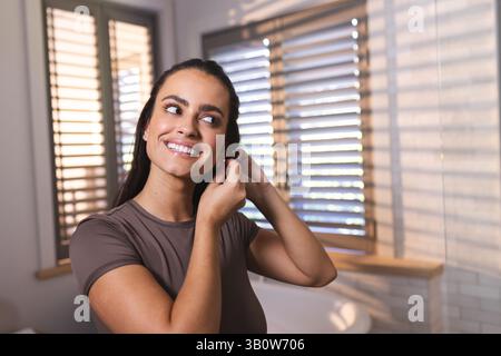 Smiling woman in her twenties adjusting pair of earrings in bathroom, with plantation shutters Stock Photo