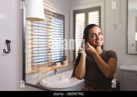 Adjusting woman putting on earring and smiling at mirror in bathroom with tub, copy space Stock Photo