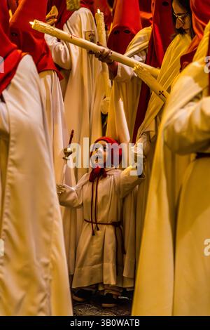A child in a white robe and red hood looks up among members of the Hermandad de San Pedro Apóstol, who take part as candle bearers in the procession on Calle Trabuco in Cuenca, Castilla-La Mancha, Spain. The brotherhood is one of 33 hermandades that stage a total of eleven processions during Cuenca's Semana Santa. Stock Photo