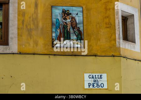 'Descendimiento' (Descent from the Cross) is the title of the painted ceramic tile picture on a yellow house wall on Calle San Juan, Cuenca, Castilla-La Mancha, Spain. It shows the body of Jesus Christ being taken down from the cross, accompanied by Mary and other figures. A street sign hangs beneath it. Stock Photo