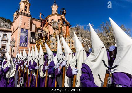 The Hermandad de Nuestro Padre Jesús Amarrado a la Columna moves with the paso of Christ's flagellation past a church façade of the Archicofradía de Paz y Caridad in Cuenca, Castilla-La Mancha, Spain. Cream-coloured hoods of the penitents fill the foreground. The brotherhood is one of 33 hermandades that stage a total of eleven processions during Cuenca's Semana Santa. Stock Photo