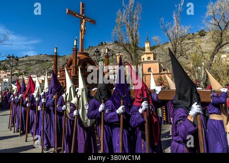 Penitents of the Archicofradía de Paz y Caridad carry a large wooden crucifix through Calle San Lázaro in Cuenca, Castilla-La Mancha, Spain. Cream, red and purple hoods mark the different hermandades affiliated with the Archicofradía. It is one of 33 hermandades that stage a total of eleven processions during Cuenca's Semana Santa. Stock Photo