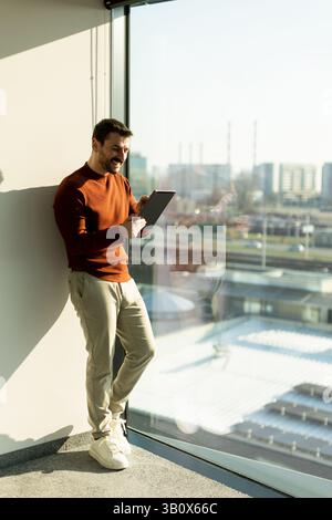 A man engages with a tablet while smiling, standing by a large window in a contemporary office. The bright sunlight highlights his thoughtful expressi Stock Photo