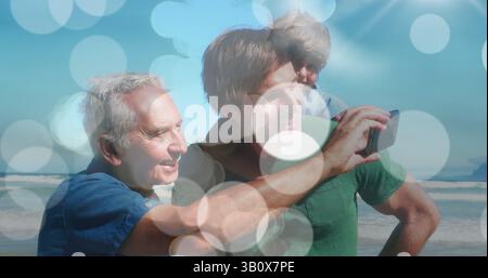 Image of bokeh over happy caucasian grandfather, father and son taking selfie on beach Stock Photo