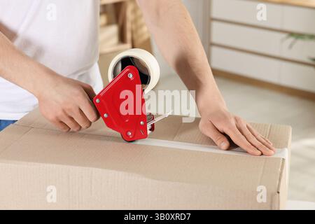 Man using dispenser with adhesive tape to seal cardboard box indoors, closeup Stock Photo
