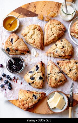 An assortment of freshly baked scones with blueberries, jam, butter, and honey on a wooden board. Stock Photo
