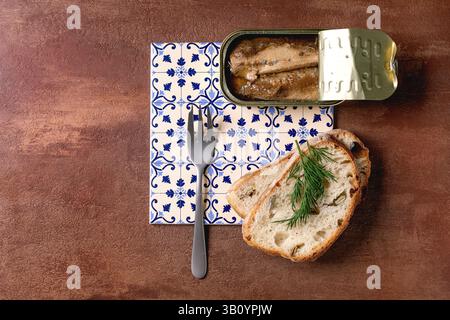 Open tin of sardine fillets in oil, served with fresh wheat bread, dill, and fork on white-and-blue patterned ceramic tiles. Textured brown background Stock Photo