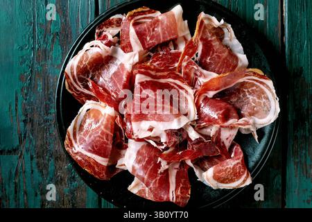 Thinly sliced Spanish ham Jamon Iberico from front pork leg paleta on black plate. Old wooden background, dark turquoise, top view. Copy space Stock Photo