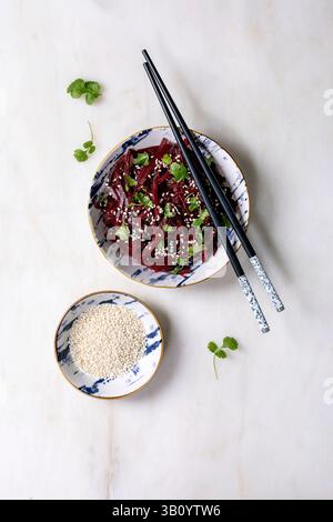 Korean-style vegan beet salad. Marinated grated beetroot with sesame seeds and coriander leaves in ceramic bowl. Nearby bowl with sesame seeds and cho Stock Photo