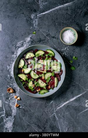 Vegan beet salad. Marinated sliced beetroot with fresh cucumber, walnuts, and coriander leaves in ceramic bowl. Nearby bowl with sea salt on black mar Stock Photo