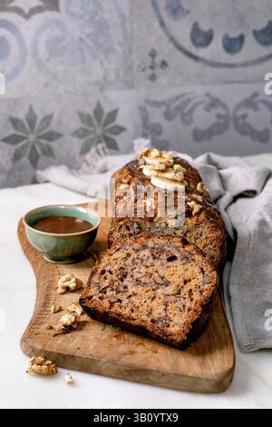 Sliced homemade banana bread with walnuts, drizzled with caramel, topped with fresh banana. Placed on wooden board with a bowl of sauce on white marbl Stock Photo
