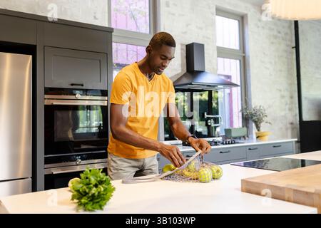African American man placing apples into reusable mesh produce bag in kitchen, with herb plant Stock Photo