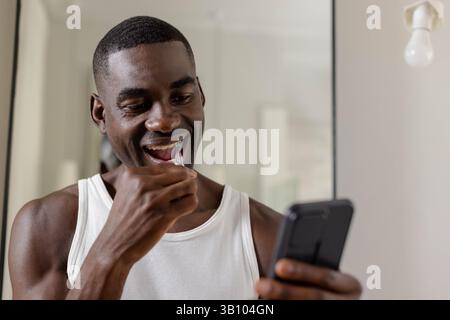 African American man brushing teeth in bathroom, holding smartphone near mirror and bulb Stock Photo