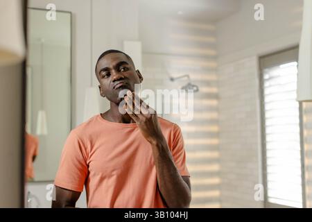 Inspecting African American man touching chin at modern bathroom mirror, with chrome showerhead Stock Photo