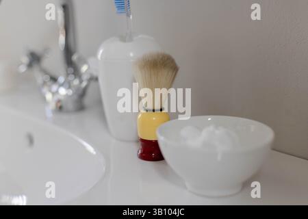 Shaving brush standing on bathroom vanity beside white sink, toothbrush holder and foam bowl Stock Photo