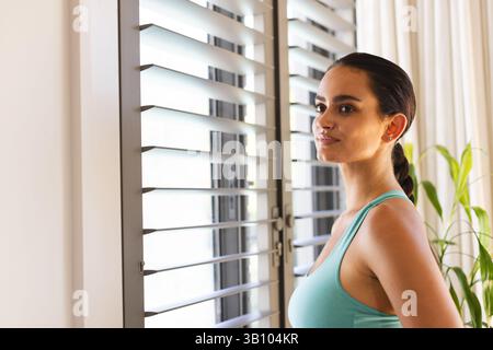 Standing woman looking outside through window blinds at home, with potted plant, copy space Stock Photo