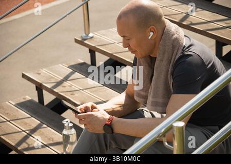 Athlete sitting on stairs and checking apps with health indicators, water balance after training. Relax after training. Man listens to music. Heat wea Stock Photo