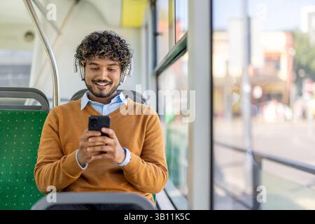 A smiling man uses his phone while riding on a bus, wearing headphones and an orange sweater Stock Photo