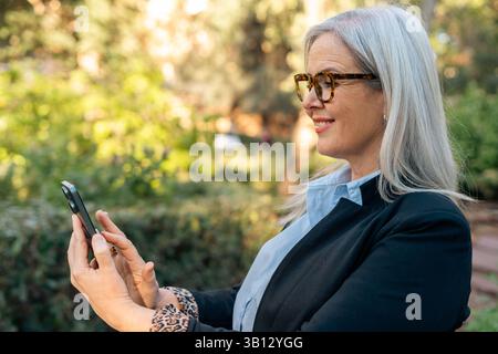 Elegant businesswoman with grey hair and glasses smiling while using a smartphone in a park, enjoying a moment of relaxation during her workday Stock Photo