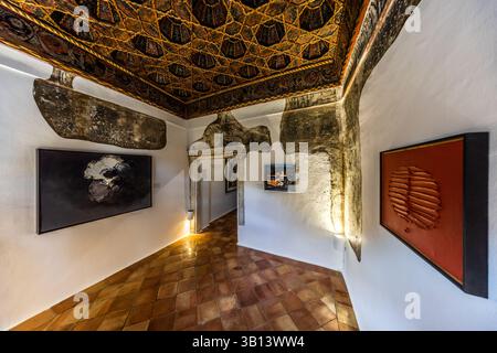 A historic ceiling painting with a geometric pattern in gold and brown tones decorates a room of the Museo de Arte Abstracto Español in Cuenca, Castilla-La Mancha, Spain. Modern paintings hang on the white walls below, including a black-and-white work and an orange relief piece. The floor is laid with brown terracotta tiles. Calle Canónigos, Cuenca, Castilla-La Mancha, Spain. Stock Photo