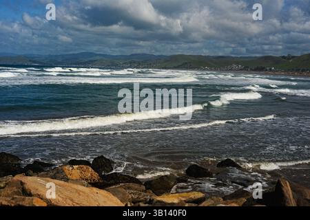 Morro Bay beach seascape, with Pacific ocean waves rolling in to beach & rock  with mountains  huge puffy white clouds & blue sky. Stock Photo