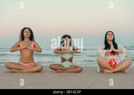 Three women sitting cross-legged on a sandy beach, practicing meditation and yoga at sunrise. The calm ocean waves and serene atmosphere enhance the p Stock Photo