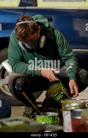 A focused artist kneels down, mixing vibrant paint colors in a container. He wears a mask and is surrounded by paint cans, showcasing a hands-on appro Stock Photo