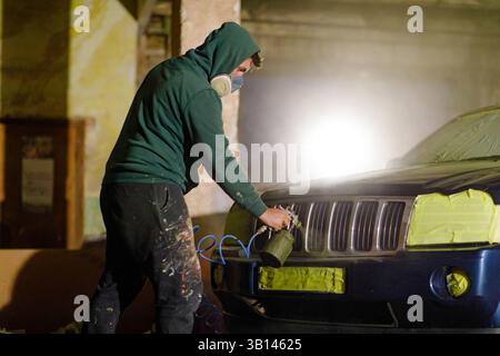 A man dressed in a green hoodie and mask carefully sprays paint on the front of a car in a dimly lit garage. The focus is on detailed work and protect Stock Photo