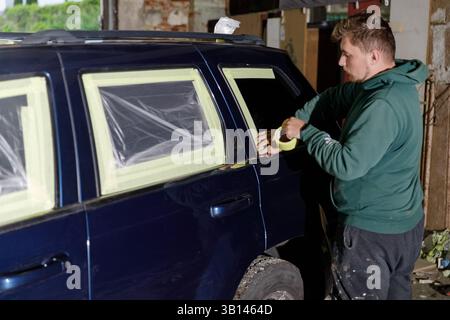 A man is getting ready to paint his car in a garage, skillfully applying masking tape around the windows to protect the areas not to be painted while Stock Photo