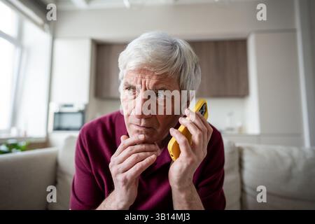 Serious elderly man appears suspicious and fearful, speaking on phone at home. Scam, fraud phobia Stock Photo