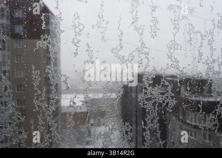a bad weather, snow patterns on glass against the background of city roofs, snowfall outside the window Stock Photo