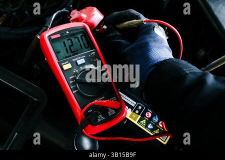 Car Battery Voltage Test, A gloved hand of technician uses a multimeter to measure the voltage of a car battery, displaying a reading of 12.70V DC Stock Photo