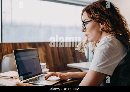 A young woman with curly red hair concentrates on her laptop in a softly lit cafe. Focused productivity, deep work sessions, digital workflow Stock Photo