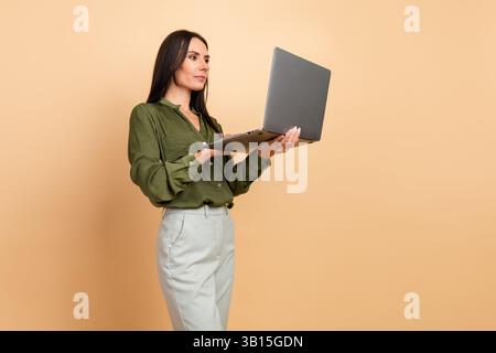 Professional woman working on a laptop while standing against a beige background in formal attire and showcasing confidence Stock Photo