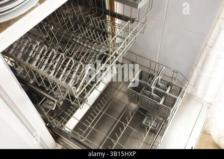 open stainless steel door of empty and clean dishwasher with pulled out rack trays with plastic cutlery basket in a white kitchen. Elevated view. Stock Photo