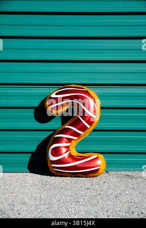 a red and white foil balloon in the shape of the number 2 leaning on a wall covered with a green corrugated metal siding Stock Photo