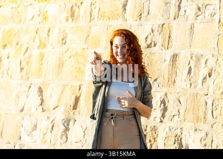 Cheerful young woman with curly red hair smiling and pointing directly at the camera with a playful expression, standing against a bright stone wall b Stock Photo