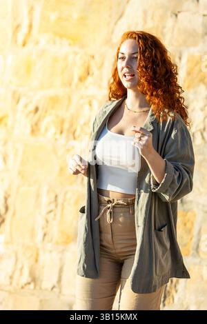 Red-haired woman with curly hair dancing with energy and a bright smile in casual outfit against a sunlit stone wall background. Lifestyle. Dance. Stock Photo