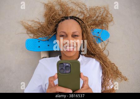 Young woman using smartphone lying on skateboard smiling Stock Photo
