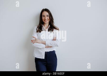Young woman stands confidently against a plain wall, wearing a white blouse and navy pants while smiling warmly Stock Photo