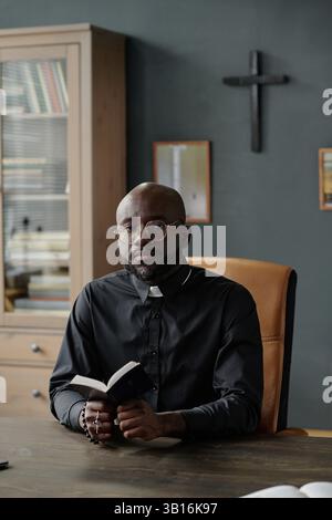 Vertical portrait of African American pastor in cassock holding bible and rosary posing for photo sitting in office Stock Photo