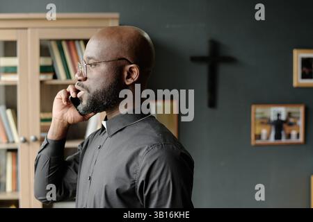 Side view of African American priest standing in office and answering phone call during work day in office Stock Photo