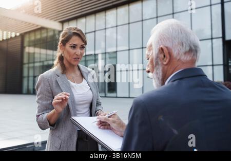 Conversation between colleagues outside office building,  woman giving instructions and senior man signing papers Stock Photo