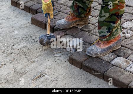 Worker using rubber mallet to set cobblestones, close-up of feet and hands on paving work. Stock Photo